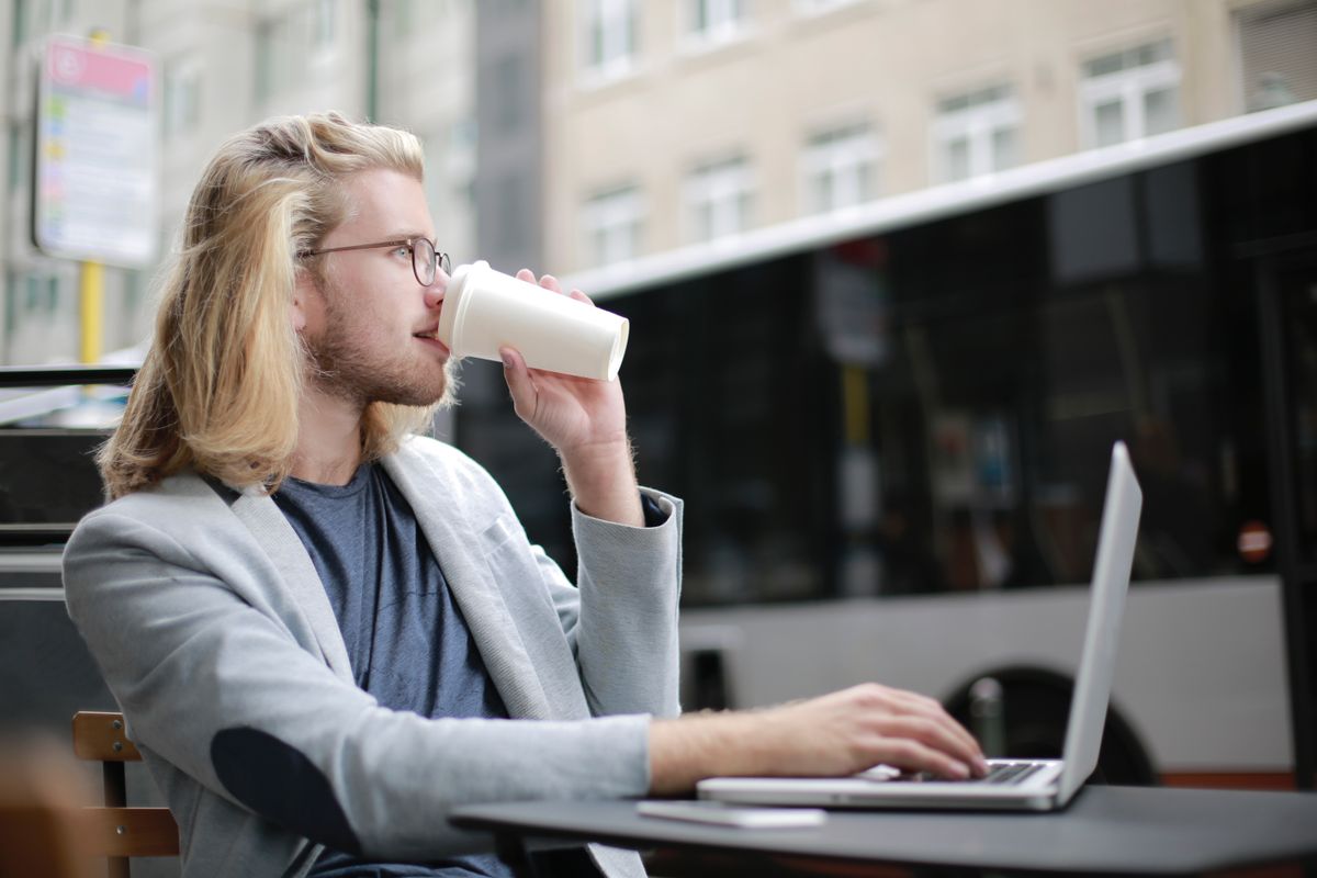 Mann med kaffekopp som jobber på laptop ved et utebord i en bygate.